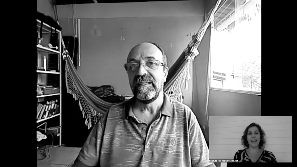 Um homem, de óculos e camisa comprida, conta sua história através da webcam do computador. Atrás dele, tem uma rede e estante de livros. No canto superior direito, tem um quadrado que mostra a webcam da entrevistadora.