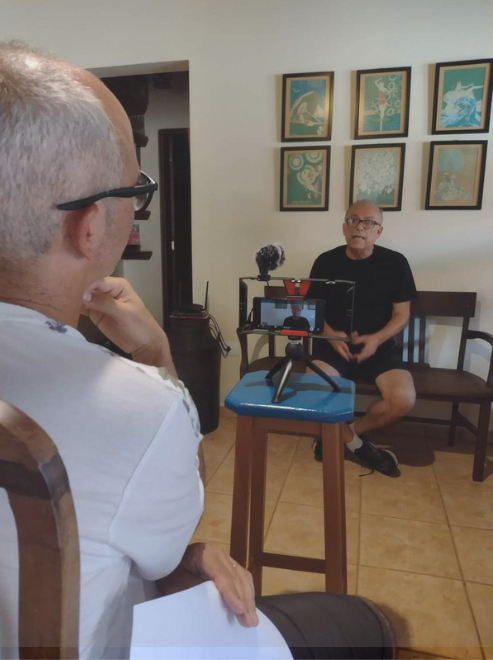 Um senhor, de cabelos brancos e óculos de grau está sentado, filando outro senhor, à sua frente. Ele usa camiseta preta, tem os cabelos grisalhos. Atrás dele, tem uma parede de quadros. A entrevista é um resultado da Formação em Memória do Museu da Pessoa.