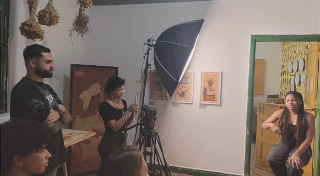 Um casal está em pé, filmando uma moça sentada. É uma entrevista do Núcleo Museu da Pessoa Vozes Daqui de Parelheiros.