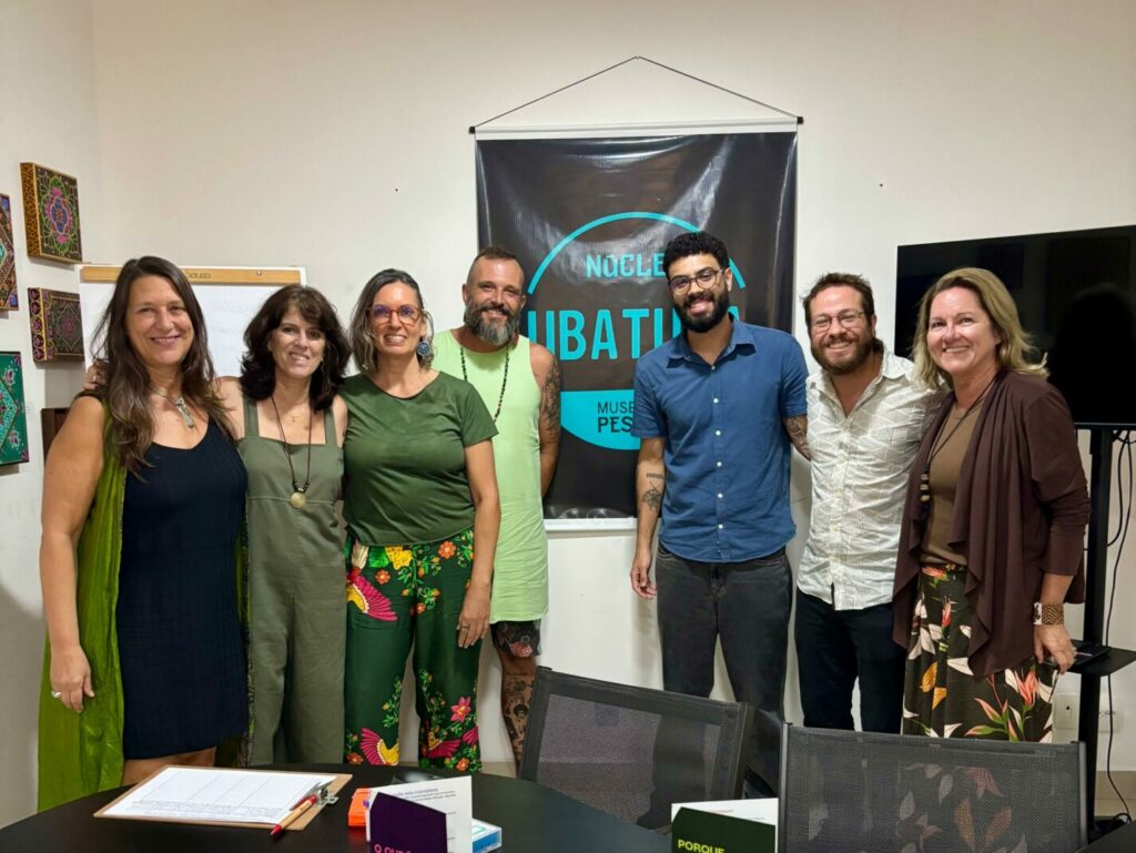 Um grupo de pessoas está em pé, em rente a um cartaz escrito Núcleo Ubatuba. Eles estão em uma sala com quadros na parede branca, um televisor ao fundo e uma mea em frente.