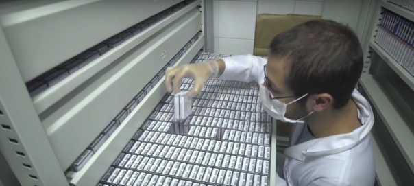 Um homem branco e loiro está usando máscara descartável e óculos. Ele está com a mão direita segurando uma acervo digital quadrado, em frente a uma gaveta.