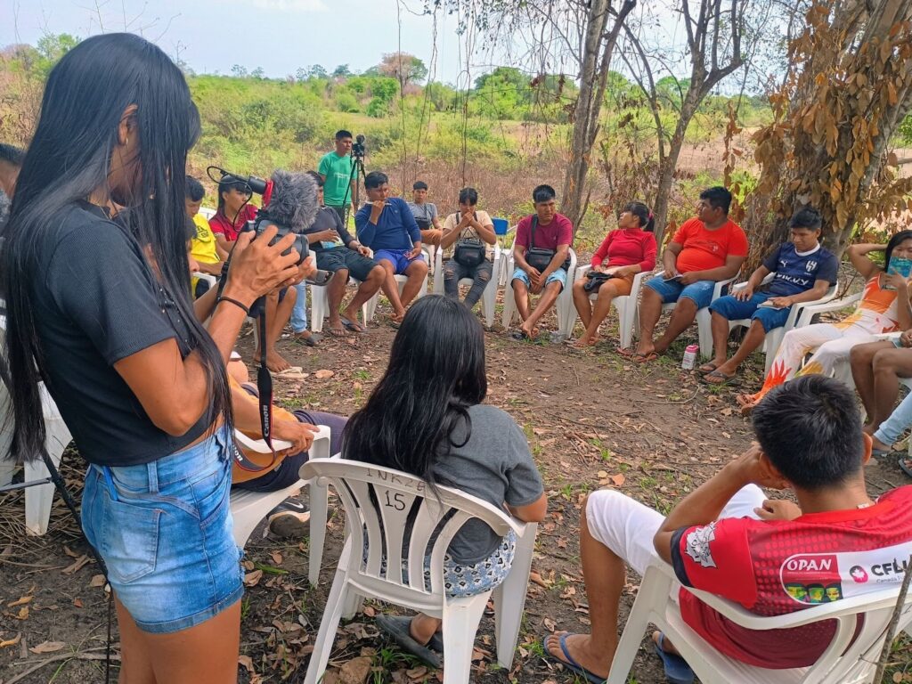 Um grupo de indígenas está sentado em cadeiras de plástico em círculo na floresta. No canto direito, uma mulher filma a atividade.