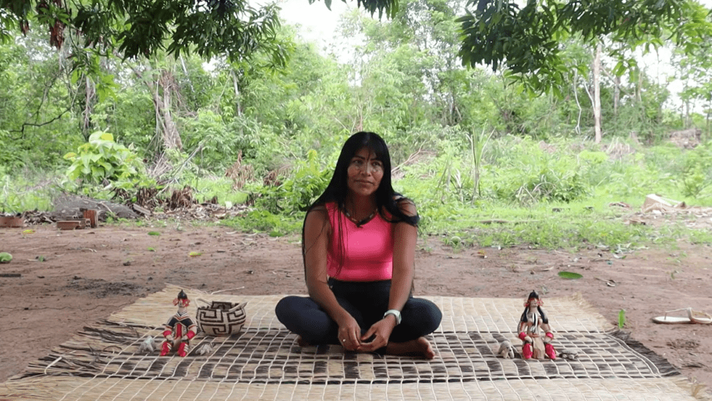 Uma mulher indígena está sentada com  as pernas cruzadas. Ela está em cima de uma rede, nela temos as bonecas karajás.