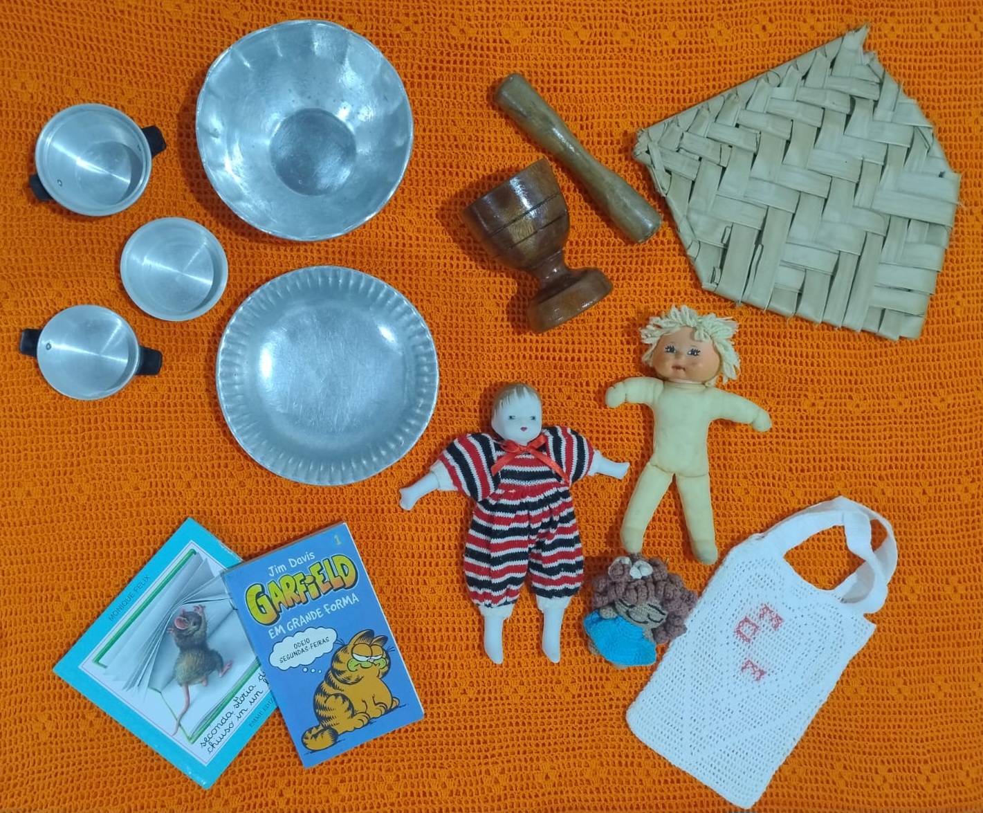 Na imagem temos peças que ajudam a contar, por meio de memórias recortes da minha vida, e da Família Marques. Peças de alumínio, produzidas a partir da reciclagem, bonecas que ganhei e fiz, livros e HQs que li, a sacolinha feita pela minha irmã, o abano usado e o pilão usados por nossa família durante muito tempo, além da colcha laranja feita pela minha mãe que serve como pano de fundo.